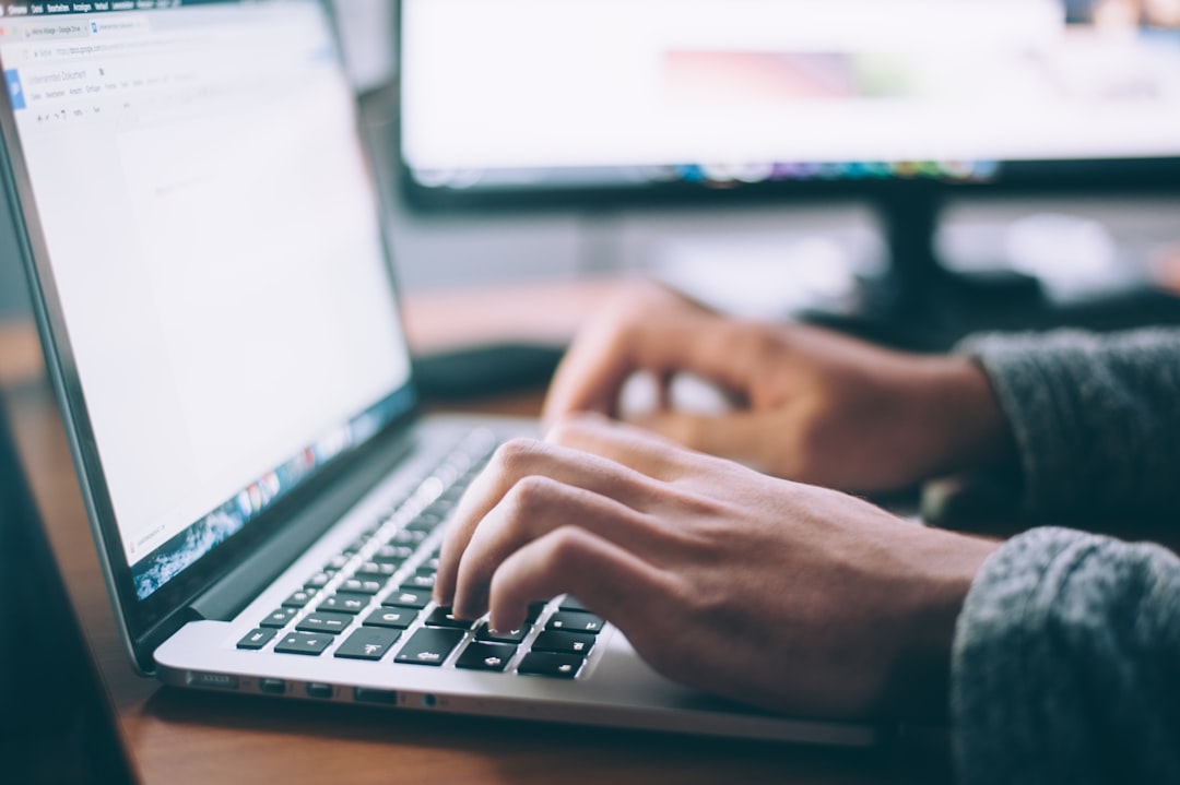 A person types on a laptop keyboard at a desk, with a monitor and blurred screen in the background, suggesting a work or study environment focused on WordPress hosting tasks.