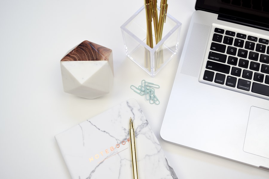A marble-patterned notebook, gold pen, paper clips, geometric container, clear pen holder with gold pens, and part of a laptop on a white desk—perfect for organizing notes on WordPress Hosting.