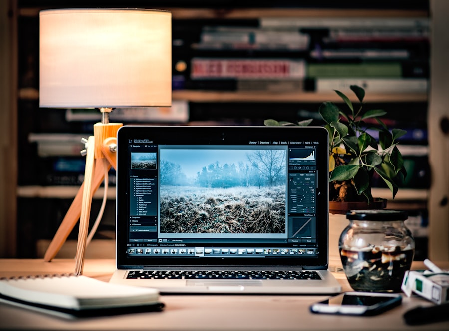 A laptop displaying photo editing software sits on a wooden desk next to a lit lamp, a potted plant, notebooks, and a glass bowl, with shelves of books and WordPress Hosting guides in the background.