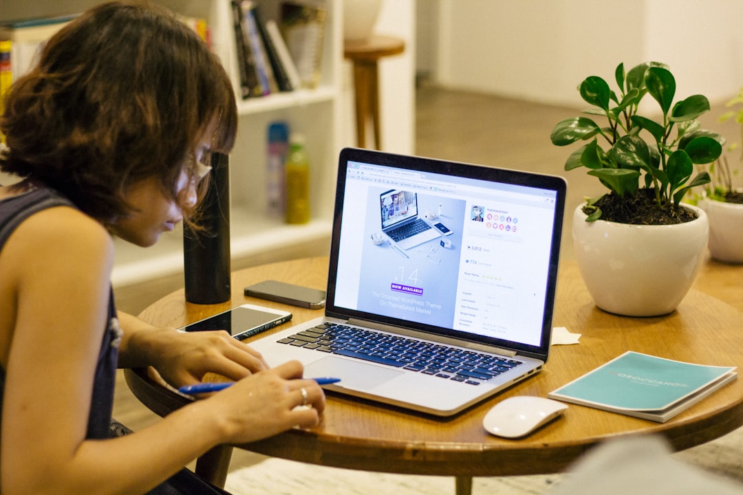 A person sits at a round wooden table, using a laptop to explore WordPress Hosting. A notebook, pen, smartphone, and potted plant are on the table. Bookshelves and another plant are visible in the background.